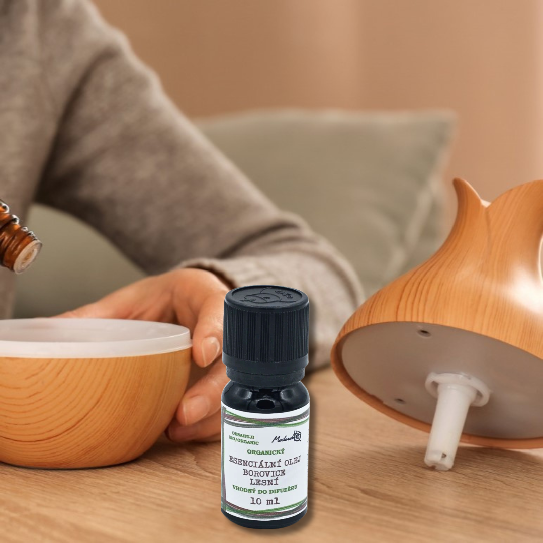 Pine essential oil bottle with a green label on a wooden table between two aroma diffusers, with a person’s hand opening one of the diffusers in the background.